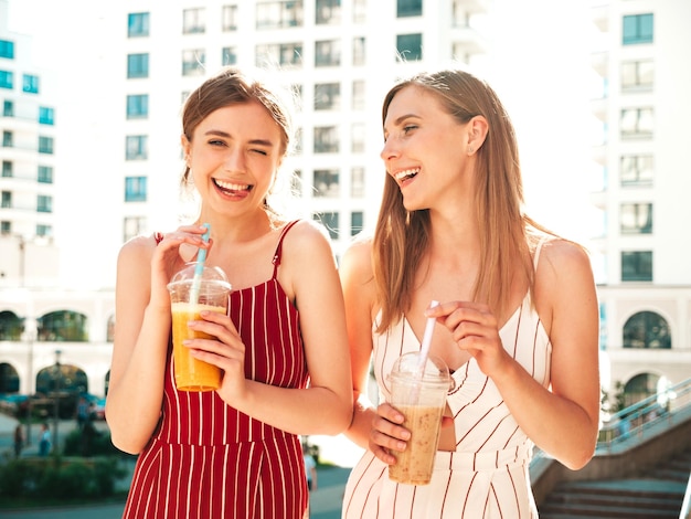 Due giovani belle donne hipster sorridenti in abiti estivi alla moda Donne spensierate in posa all'apertoModelli positivi che tengono e bevono cocktail fresco frullato in bicchiere di plastica con cannuccia