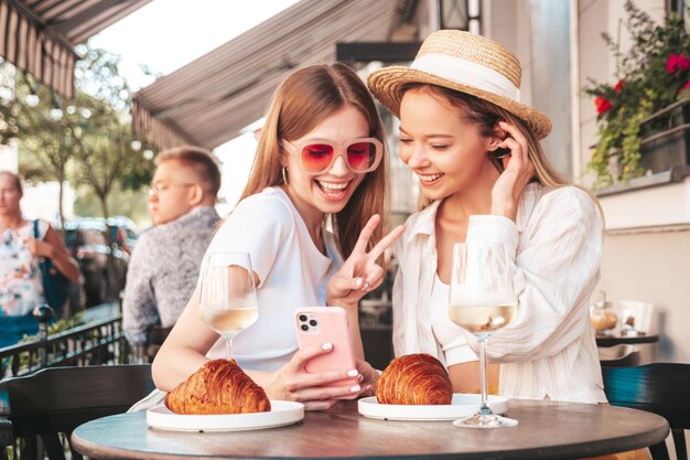 Due giovani belle donne hipster sorridenti in abiti estivi alla moda Donne spensierate sedute al caffè sulla veranda in stradaModelli positivi che bevono vino biancoMangiare croissant Guardando lo schermo del telefono