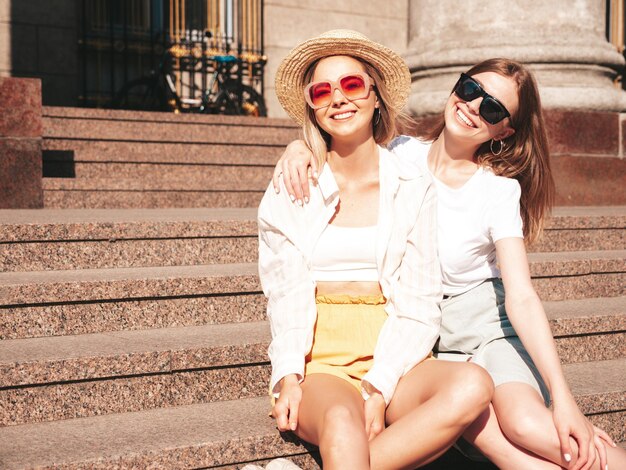 Due giovani belle donne hipster sorridenti in abiti estivi alla moda Donne spensierate sexy che posano per strada con cappello Modelli puri positivi che si divertono al tramonto