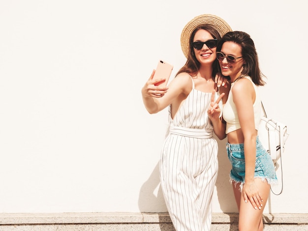 Due giovani belle donne hipster sorridenti in abiti estivi alla moda Donne spensierate sexy in posa per strada vicino al muro bianco con cappello Modelli puri positivi che si divertono al tramonto prendendo selfie