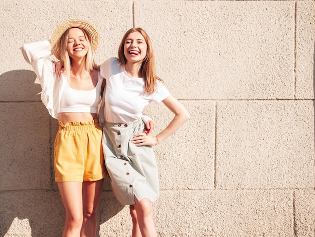 Due giovani belle donne hipster sorridenti in abiti estivi alla moda Donne spensierate sexy in posa per strada vicino al muro bianco con cappello Modelli puri positivi che si divertono al tramonto