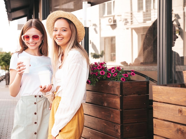 Due giovani belle donne hipster sorridenti in abiti estivi alla modaDonne spensierate sexy che posano per strada Modelli puri positivi che si divertono al tramonto Bevono caffè o tè in un bicchiere di plastica