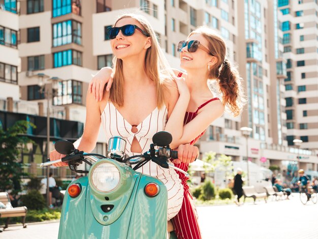 Due giovani belle donne hipster sorridenti in tuta alla modaDonne spensierate sexy che guidano una moto retrò sullo sfondo della stradaModelli positivi che si divertono a cavalcare un classico scooter italiano in occhiali