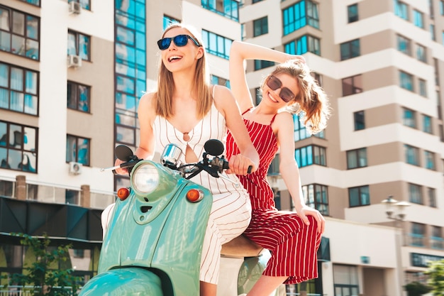 Due giovani belle donne hipster sorridenti in tuta alla modaDonne spensierate sexy che guidano una moto retrò sullo sfondo della stradaModelli positivi che si divertono a cavalcare un classico scooter italiano in occhiali