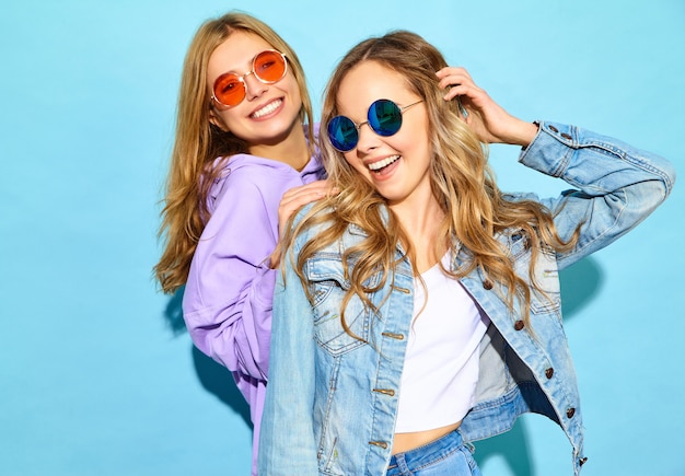 Due giovani belle donne sorridenti bionde dei pantaloni a vita bassa in vestiti d'avanguardia di estate. Donne spensierate sexy che posano vicino alla parete blu in occhiali da sole. I modelli positivi impazziscono