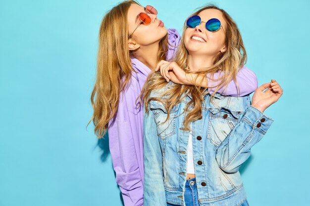 Due giovani belle donne sorridenti bionde dei pantaloni a vita bassa in vestiti d'avanguardia di estate. Donne spensierate sexy che posano vicino alla parete blu in occhiali da sole. Modelli positivi che impazziscono e si abbracciano