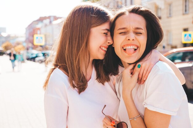 Due giovani belle donne sorridenti dei pantaloni a vita bassa in vestiti estivi alla moda. Donne spensierate sexy che posano nella via. Modelle pure positive che si divertono al tramonto, si abbracciano e impazziscono. Felice e allegro