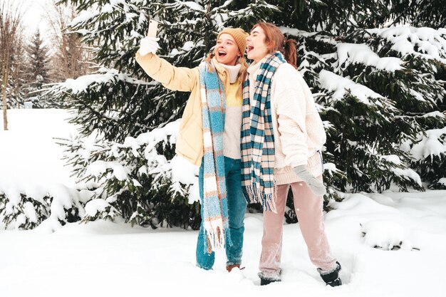 Due giovani belle donne sorridenti hipster in vestiti caldi alla moda e sciarpe. Donne spensierate in posa in strada nel parco. Modelli puri positivi che si divertono nella neve. Godersi i momenti invernali.Prendere selfie