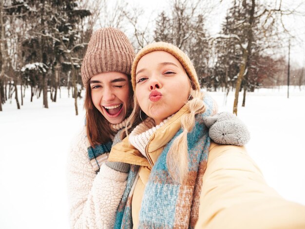 Due giovani belle donne sorridenti hipster in vestiti caldi alla moda e sciarpe. Donne spensierate in posa in strada nel parco. Modelli puri positivi che si divertono nella neve. Godersi i momenti invernali.Prendere selfie