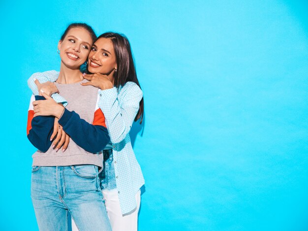 Due giovani belle ragazze bionde sorridenti dei pantaloni a vita bassa in vestiti variopinti della maglietta di estate alla moda. Donne spensierate sexy che posano vicino alla parete blu. Modelli positivi che si divertono