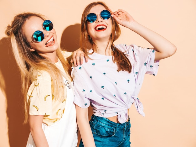 Due giovani belle ragazze bionde sorridenti dei pantaloni a vita bassa in vestiti variopinti della maglietta di estate alla moda. e mostrando la lingua