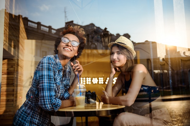 Due giovani belle ragazze che sorridono, parlando, riposando nella caffetteria. Sparato dall'esterno.