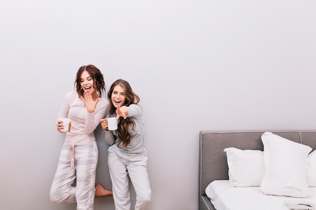 Due giovani belle ragazze in pigiama di notte con le tazze che hanno divertimento nella camera da letto sul muro grigio. Sembrano divertiti e sorridenti.