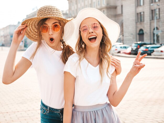 Due giovani belle ragazze sorridenti bionde dei pantaloni a vita bassa in vestiti bianchi alla moda della maglietta di estate. Donne colpite sexy che posano nella via. Modelli sorpresi che si divertono in occhiali da sole e cappello. Mostra il segno di pace