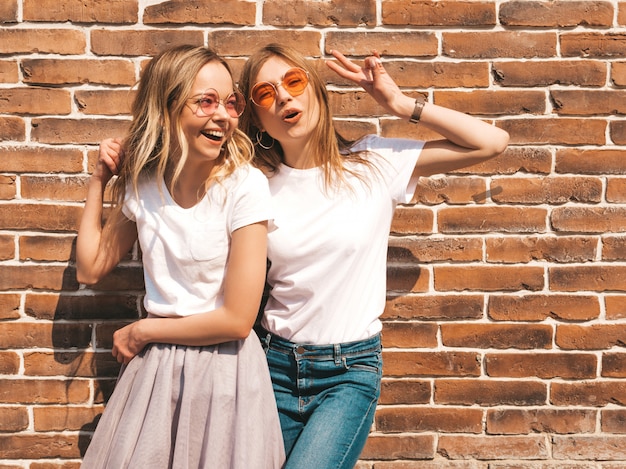 Due giovani belle ragazze sorridenti bionde dei pantaloni a vita bassa in vestiti bianchi alla moda della maglietta di estate. . Modelli positivi che si divertono in occhiali da sole. Mostra il segno di pace