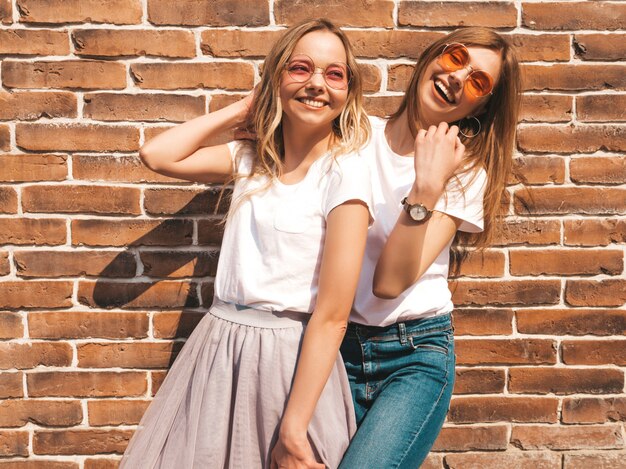 Due giovani belle ragazze sorridenti bionde dei pantaloni a vita bassa in vestiti bianchi alla moda della maglietta di estate. . Modelli positivi che si divertono in occhiali da sole