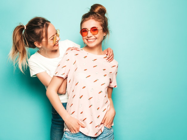 Due giovani belle ragazze sorridenti bionde dei pantaloni a vita bassa in vestiti d'avanguardia dei jeans dei pantaloni a vita bassa dell'estate. Donne spensierate sexy che posano vicino alla parete blu. Modelli trendy e positivi che si divertono con gli occhiali da sole