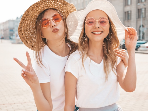 Due giovani belle ragazze sorridenti bionde dei pantaloni a vita bassa in vestiti d'avanguardia di estate. Mostra il segno di pace