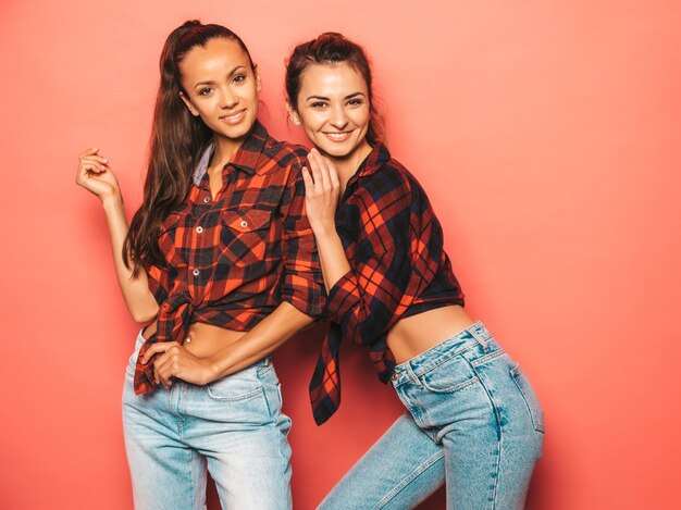 Due giovani belle ragazze sorridenti dei pantaloni a vita bassa del brunette in vestiti a quadretti simili d'avanguardia della camicia e dei jeans Donne spensierate sexy che posano vicino alla parete rosa in studio Divertimento dei modelli positivi