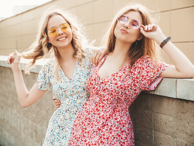 Due giovani belle ragazze sorridenti dei pantaloni a vita bassa in prendisole estive d'avanguardia Donne sexy spensierate che posano sui precedenti della via in occhiali da sole. Modelli positivi che si divertono e si abbracciano