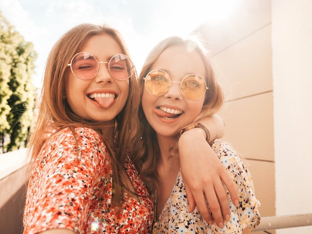 Due giovani belle ragazze sorridenti dei pantaloni a vita bassa in prendisole estive d'avanguardia Donne sexy spensierate che posano sui precedenti della via in occhiali da sole. Scattano selfie selfie su smartphone al tramonto