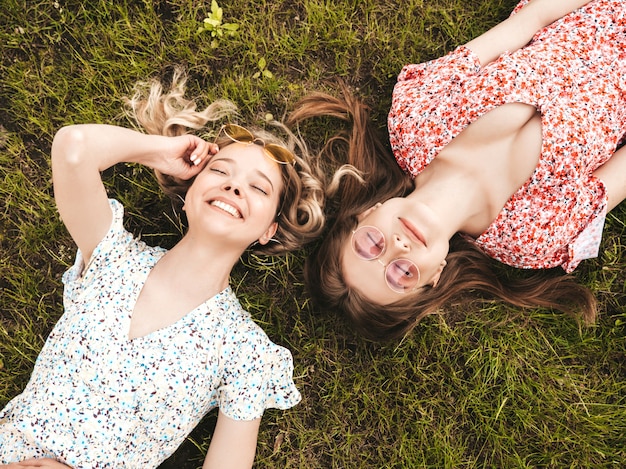 Due giovani belle ragazze sorridenti dei pantaloni a vita bassa in prendisole estive d'avanguardia Donne spensierate sexy che si trovano sull'erba verde in occhiali da sole Divertimento dei modelli positivi Vista superiore