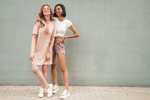 Due giovani belle ragazze sorridenti dei pantaloni a vita bassa in vestiti alla moda di estate. Donne spensierate sexy che posano vicino alla parete nella via in occhiali da sole. Modelli positivi che si divertono e si abbracciano