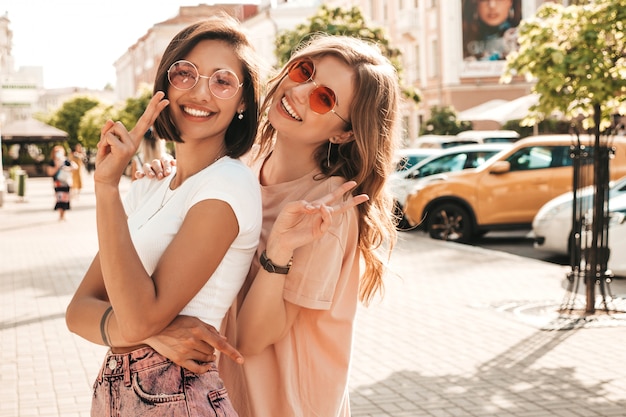 Due giovani belle ragazze sorridenti dei pantaloni a vita bassa in vestiti d'avanguardia di estate Donne spensierate sexy che posano sui precedenti della via in occhiali da sole. Modelle positive che si divertono e si abbracciano. Impazziscono