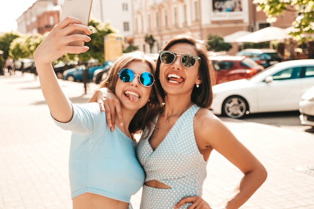 Due giovani belle ragazze sorridenti dei pantaloni a vita bassa in vestiti d'avanguardia di estate Donne spensierate sexy che posano sui precedenti della via in occhiali da sole. Scattano selfie selfie su smartphone al tramonto