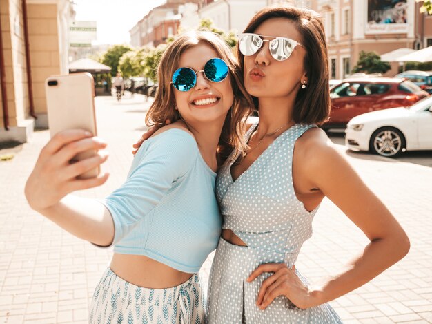 Due giovani belle ragazze sorridenti dei pantaloni a vita bassa in vestiti d'avanguardia di estate Donne spensierate sexy che posano sui precedenti della via in occhiali da sole. Scattano selfie selfie su smartphone al tramonto