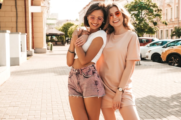 Due giovani belle ragazze sorridenti dei pantaloni a vita bassa in vestiti d'avanguardia di estate Donne spensierate sexy che posano sul fondo della via in occhiali da sole. Modelle positive che si divertono e impazziscono