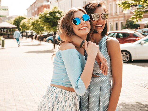 Due giovani belle ragazze sorridenti dei pantaloni a vita bassa in vestiti d'avanguardia di estate Donne spensierate sexy che posano sul fondo della via in occhiali da sole. Modelli positivi che si divertono e si abbracciano