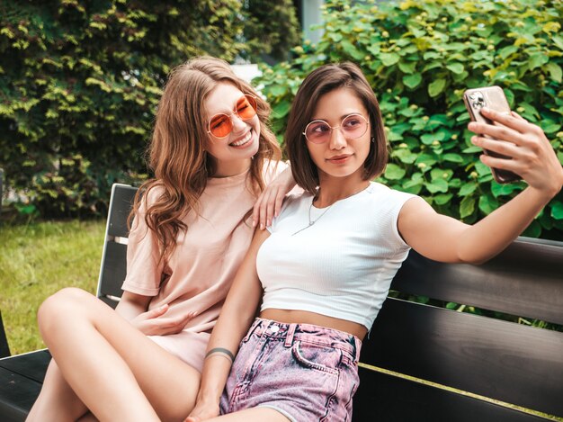 Due giovani belle ragazze sorridenti dei pantaloni a vita bassa in vestiti estivi d'avanguardia Donne spensierate sexy che si siedono sul banco sulla via in occhiali da sole. Stanno scattando foto di autoritratto con selfie su smartphone