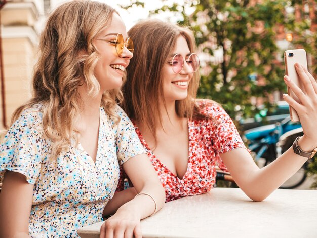 Due giovani belle ragazze sorridenti dei pantaloni a vita bassa nelle prendisole estive d'avanguardia Donne libere che chiacchierano nel caffè della veranda sui precedenti della via Modelli positivi divertendosi e prendendo selfie sullo smartphone