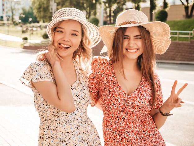 Due giovani belle ragazze sorridenti dei pantaloni a vita bassa nelle prendisole estive d'avanguardia Donne spensierate sexy che posano sui precedenti della via in cappelli. Modelli positivi che si divertono e abbracciano. Mostrano segno di pace e lingua