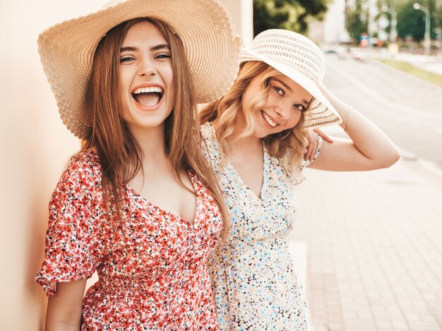 Due giovani belle ragazze sorridenti dei pantaloni a vita bassa nelle prendisole estive d'avanguardia Donne spensierate sexy che posano sui precedenti della via in cappelli. Modelli positivi che si divertono e si abbracciano