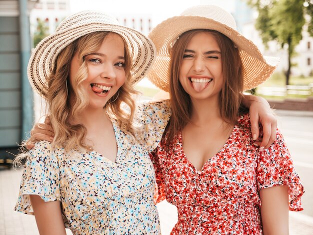 Due giovani belle ragazze sorridenti dei pantaloni a vita bassa nelle prendisole estive d'avanguardia Donne spensierate sexy che posano sul fondo della via in cappelli. Modelle positive che si divertono e si abbracciano. Mostrano lingue