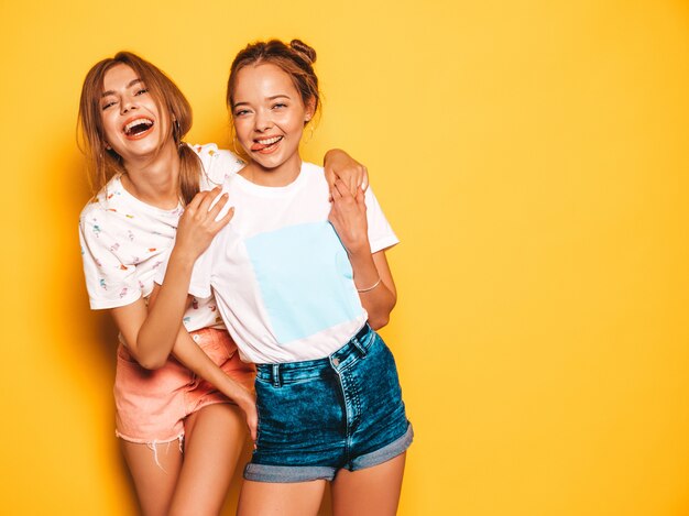 Due giovani belle ragazze sorridenti hipster in abiti estivi alla moda. Donne spensierate sexy che posano vicino alla parete gialla. Modelle positive che impazziscono e si divertono