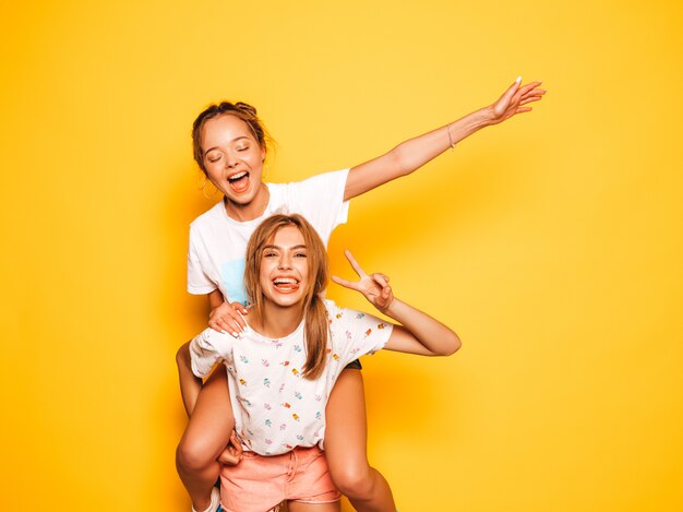 Due giovani belle ragazze sorridenti hipster in abiti estivi alla moda. Donne spensierate sexy che posano vicino alla parete gialla. Modello che si siede sulla schiena dell'amica e che solleva le mani