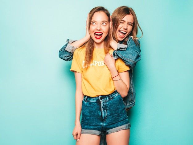 Due giovani belle ragazze sorridenti hipster in giacca estiva t-shirt e jeans giallo alla moda. Donne spensierate sexy che posano vicino alla parete blu. Modelli alla moda e positivi che si divertono