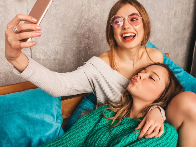 Due giovani belle ragazze sorridenti splendide in abiti estivi alla moda. Donne spensierate sexy che posano nell'interno e che prendono selfie. Modelli positivi che si divertono con lo smartphone.