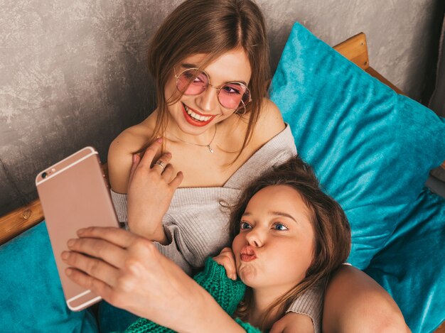 Due giovani belle ragazze sorridenti splendide in abiti estivi alla moda. Donne spensierate sexy che posano nell'interno e che prendono selfie. Modelli positivi che si divertono con lo smartphone.