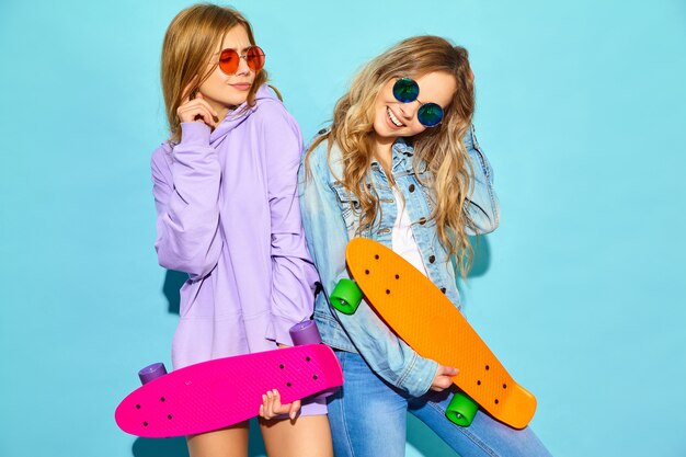 Due giovani donne bionde sorridenti alla moda con i pattini del penny. donne in abiti sportivi estate hipster in posa vicino al muro blu. Modelli positivi