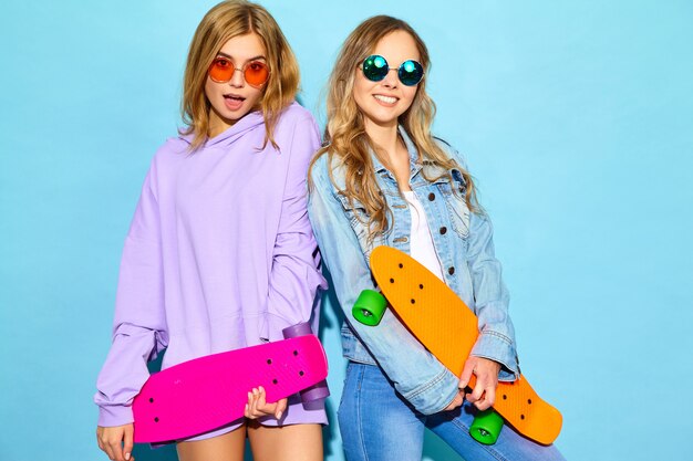 Due giovani donne bionde sorridenti alla moda con i pattini del penny. Modelli in abiti sportivi hipster estate in posa vicino alla parete blu. Donne positive che impazziscono