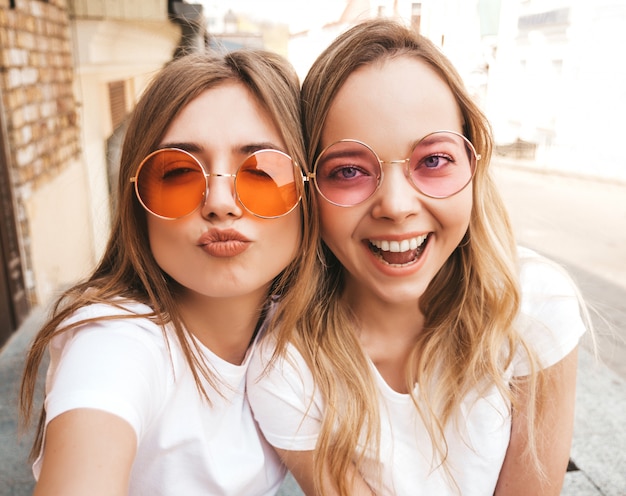 Due giovani donne bionde sorridenti dei pantaloni a vita bassa in maglietta bianca di estate. Ragazze che prendono le foto dell'autoritratto del selfie sullo smartphone. Faccia femminile che fa la faccia di anatra