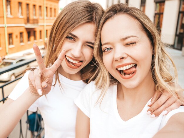 Due giovani donne bionde sorridenti dei pantaloni a vita bassa in vestiti bianchi della maglietta di estate. Ragazze che prendono le foto dell'autoritratto del selfie sullo smartphone. .Female mostra segno di pace e lingua