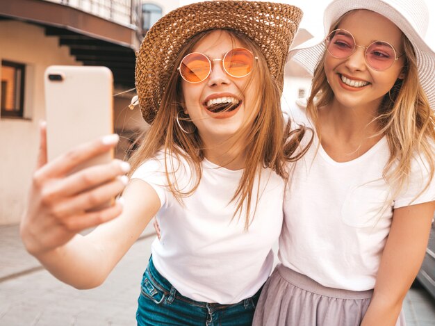 Due giovani donne bionde sorridenti dei pantaloni a vita bassa in vestiti bianchi della maglietta di estate. Ragazze che prendono le foto dell'autoritratto del selfie sullo smartphone.