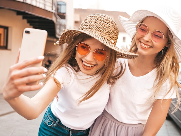 Due giovani donne bionde sorridenti dei pantaloni a vita bassa in vestiti bianchi della maglietta di estate. Ragazze che prendono le foto dell'autoritratto del selfie sullo smartphone.