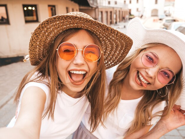 Due giovani donne bionde sorridenti dei pantaloni a vita bassa in vestiti bianchi della maglietta di estate. Ragazze che prendono le foto dell'autoritratto del selfie sullo smartphone.