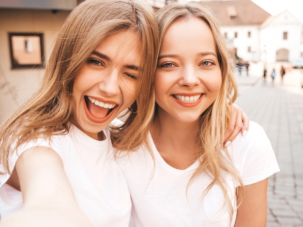 Due giovani donne bionde sorridenti dei pantaloni a vita bassa in vestiti bianchi della maglietta di estate. Ragazze che prendono le foto dell'autoritratto del selfie sullo smartphone.